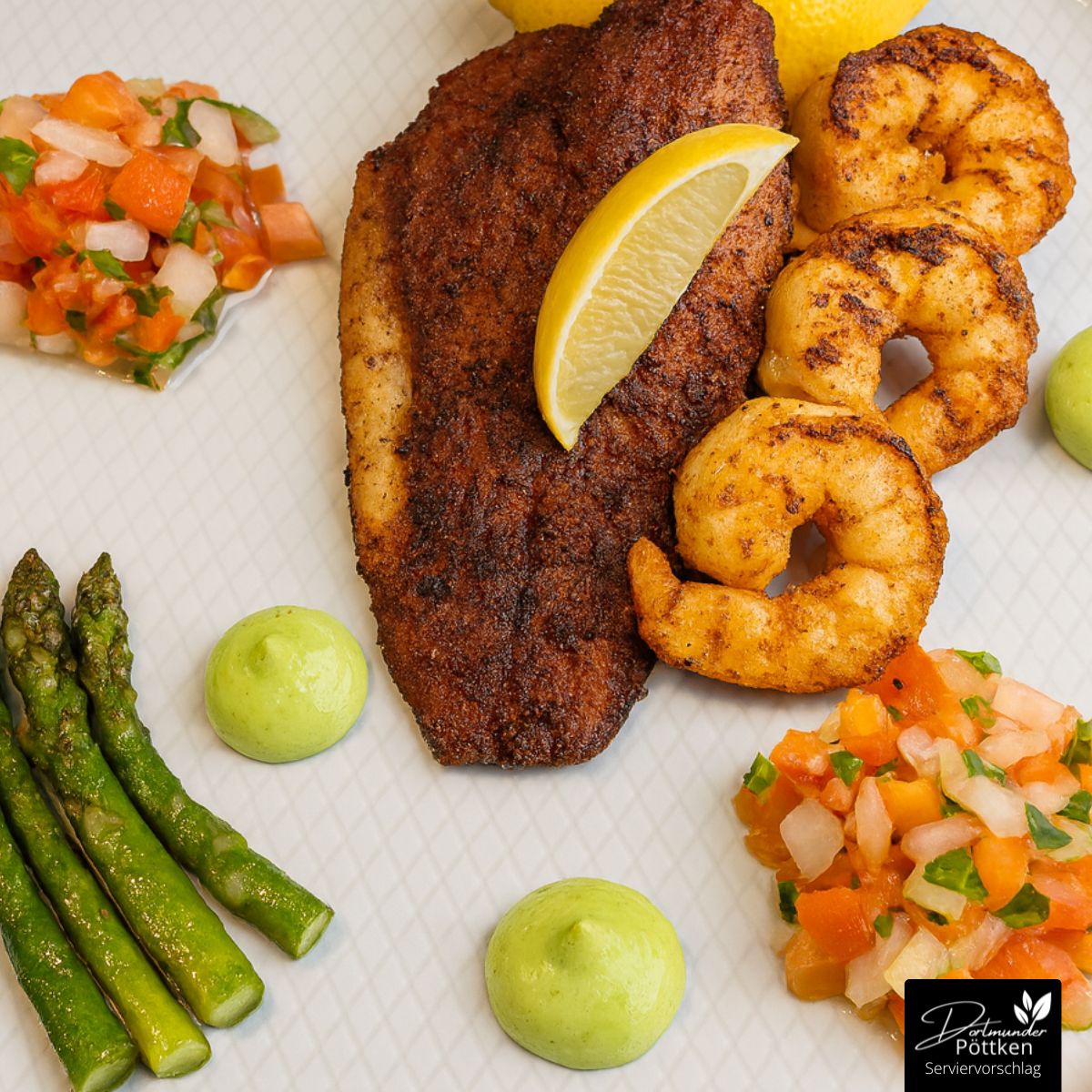 Blackened Fisch mit Garnelen, Spargel und Avocadocreme Ein weißer Teller mit Blackened Fischfilet, gegrillten Garnelen, grünem Spargel, Avocadocreme und frischem Tomaten-Salsa, garniert mit einer Zitronenscheibe.