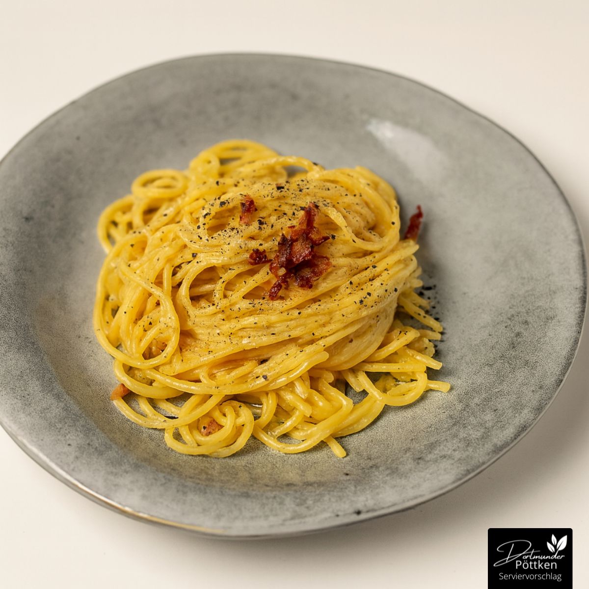 Spaghetti Carbonara auf grauem Teller, garniert mit schwarzem Pfeffer und knusprigen Speckstücken