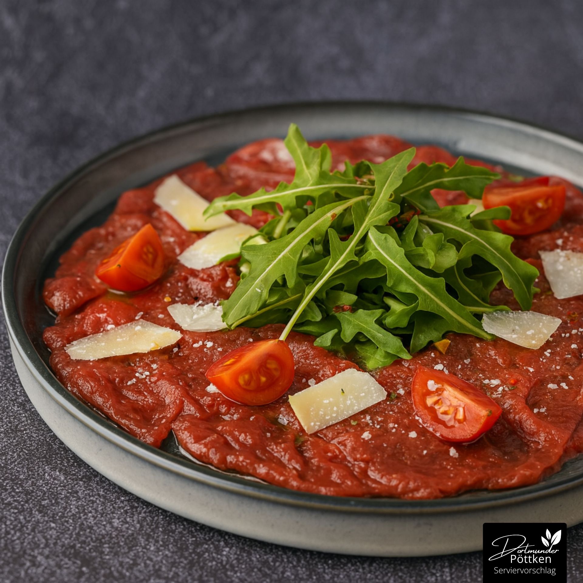 Rindercarpaccio mit Rucola, Parmesan, Fleur de Sel und Kirschtomaten auf grauem Teller angerichtet.